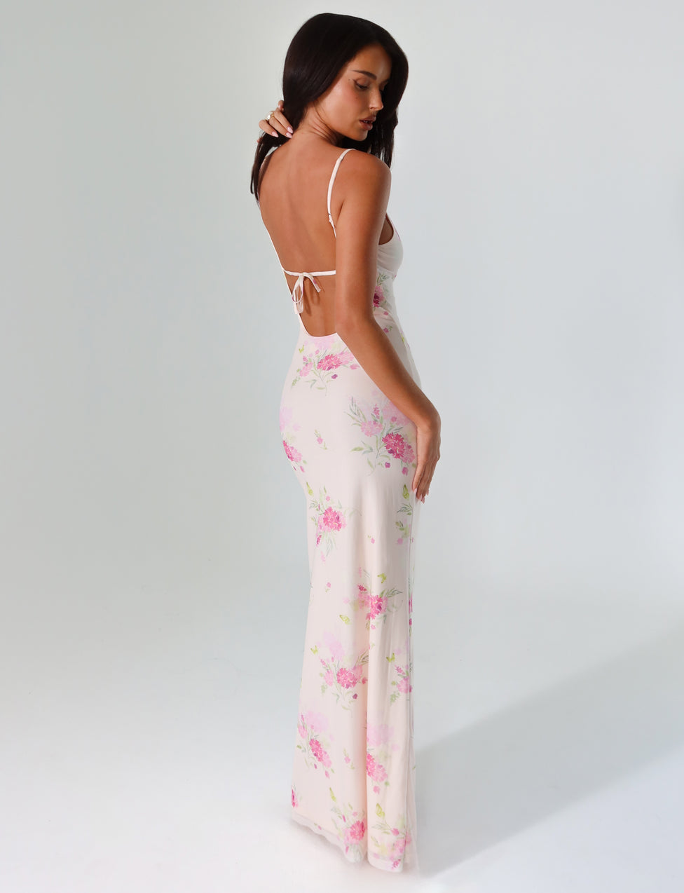 A woman stands against a plain background wearing the OSIRIS Dress by TIGERMIST. This long, open-back dress showcases a deep V neckline and features a watercolor floral print with pink and purple flowers on a white base. She is slightly turned to the side, with her arms by her sides, looking downward.