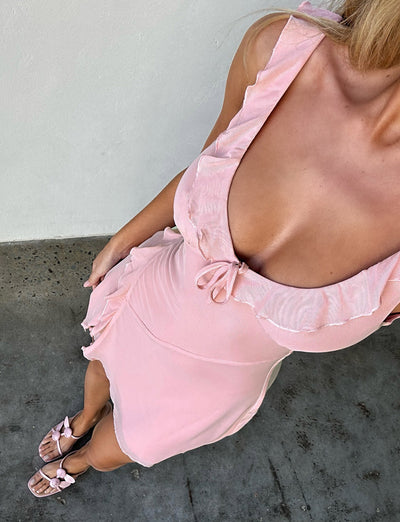 A person is wearing the TIGERMIST IMRA DRESS in dusty pink, featuring ruffled edges and a bow at the neckline. They are wearing sandals with bow accents and standing on a concrete surface. The image is taken from a high angle, focusing on their upper body and attire.