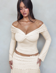 A woman with long brown hair wears the TIGERMIST DENALI LONG SLEEVE TOP in White : Bone, paired with a fitted cream skirt, standing against a light gray background and looking down with her hands on her hips.