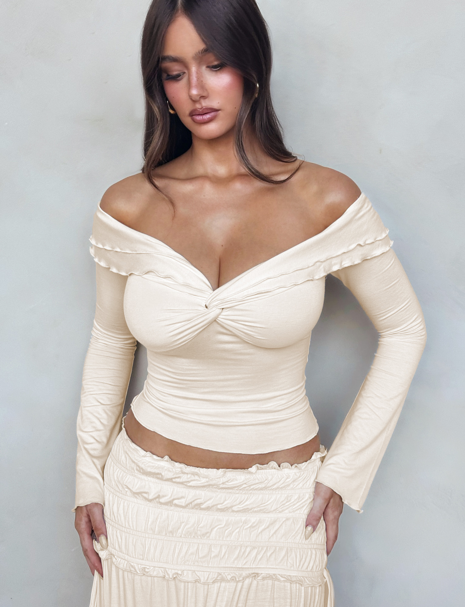 A woman with long brown hair wears the TIGERMIST DENALI LONG SLEEVE TOP in White : Bone, paired with a fitted cream skirt, standing against a light gray background and looking down with her hands on her hips.