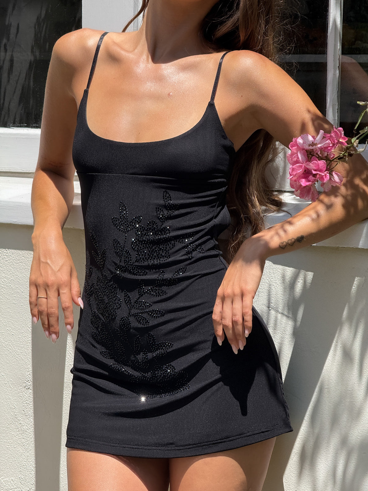 A woman stands by a white window in the TIGERMIST OCTAVIA MINI DRESS - BLACK, featuring floral embellishments and adjustable spaghetti straps. Sunlight highlights her skin and long wavy hair as she holds a pink flower, her face partially hidden.