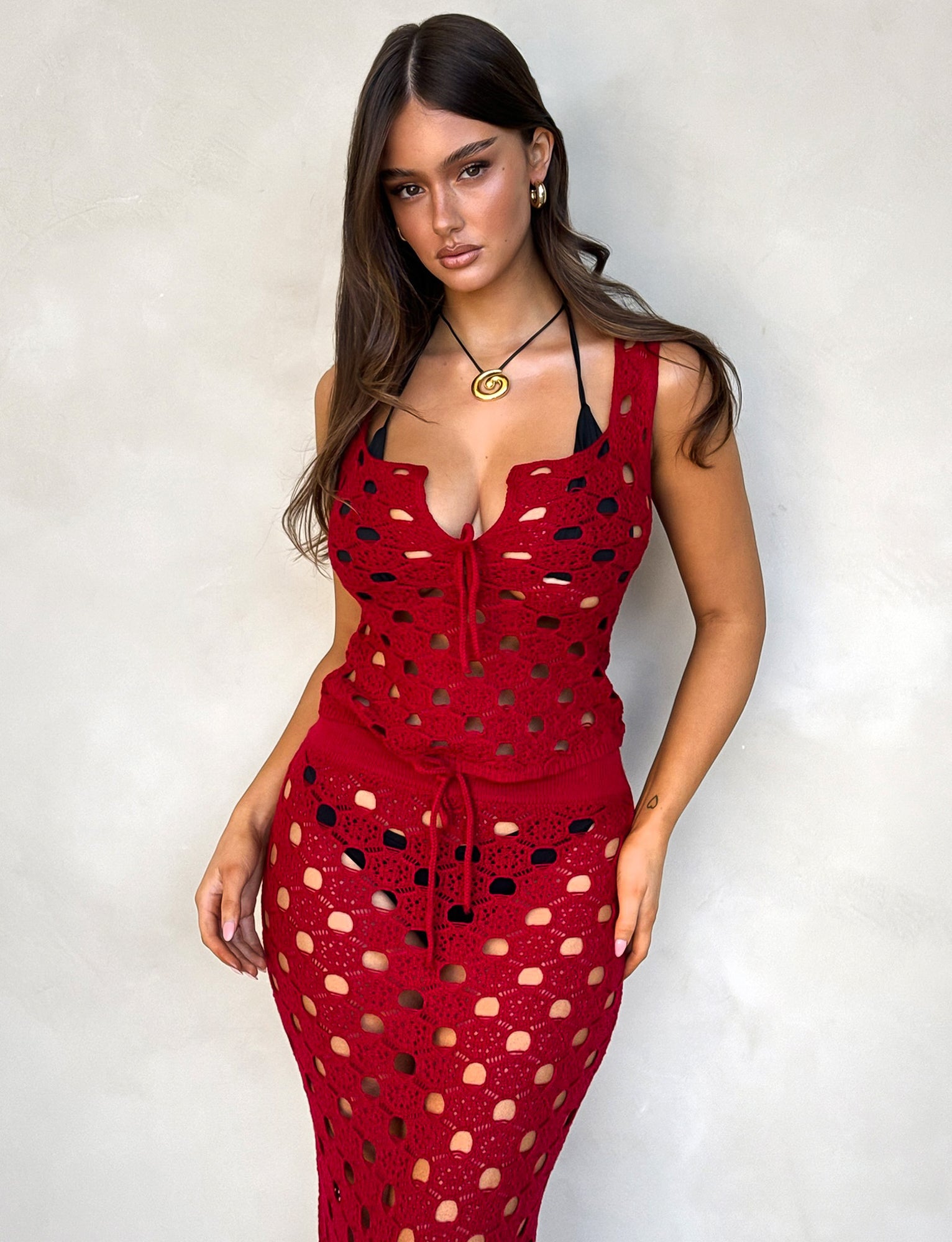 A woman with long brown hair wears the KINCADE TOP - RED: CHERRY RED from TIGERMIST, featuring a crochet-style design with circular cut-outs that reveal a black bikini underneath. She stands against a plain, light-colored background, accessorized with a gold necklace and earrings, displaying a calm, confident expression.