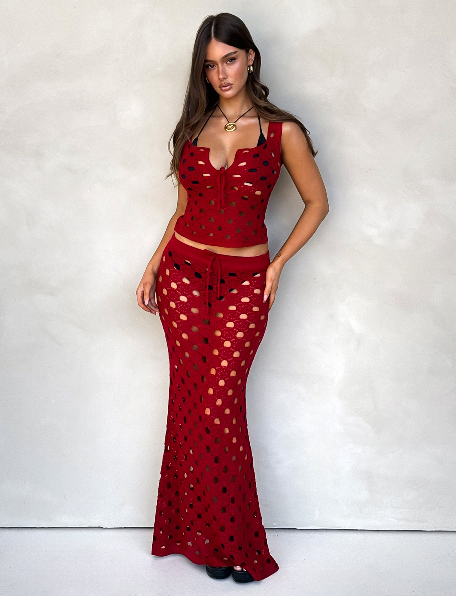 A woman stands against a plain backdrop, exuding elegance in a matching red crochet top and the Kincade Skirt in Cherry Red from Tiger Mist. Her long, wavy hair is parted in the middle, and she's accessorized with gold jewelry. She has a neutral expression, one hand on her hip and the other at her side.