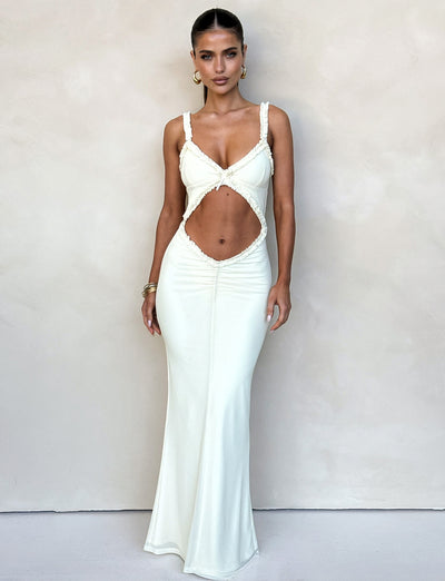 A woman stands against a plain beige wall, wearing the RAYLA DRESS - WHITE : CREAM from TIGERMIST, a long, fitted dress featuring a cutout section in the midriff area. Her hair is pulled back and she accessorizes with hoop earrings and bracelets. She looks directly at the camera with a serious expression.