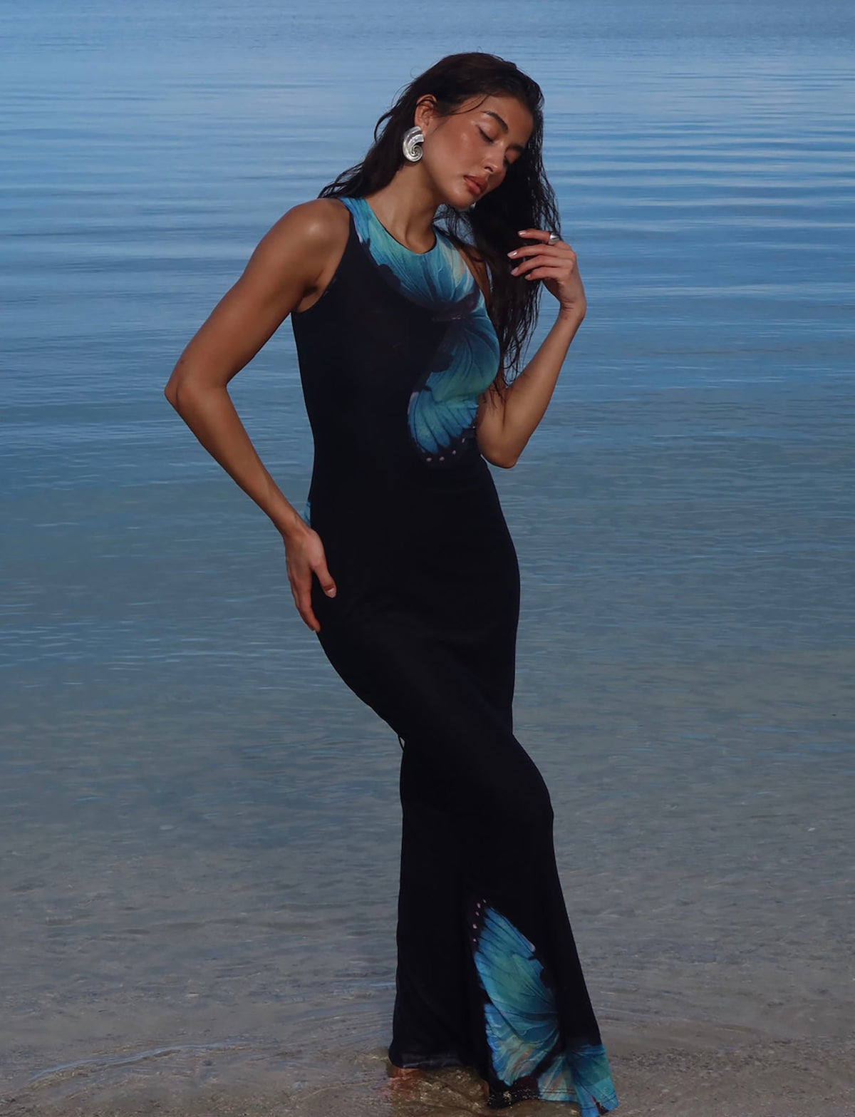 A woman in the TIGERMIST ZAHRAA DRESS - BLACK : BUTTERFLY stands in shallow water. She gently touches her wet hair, looking down with a serene expression. The calm sea in the background merges with the clear sky, creating a tranquil scene.