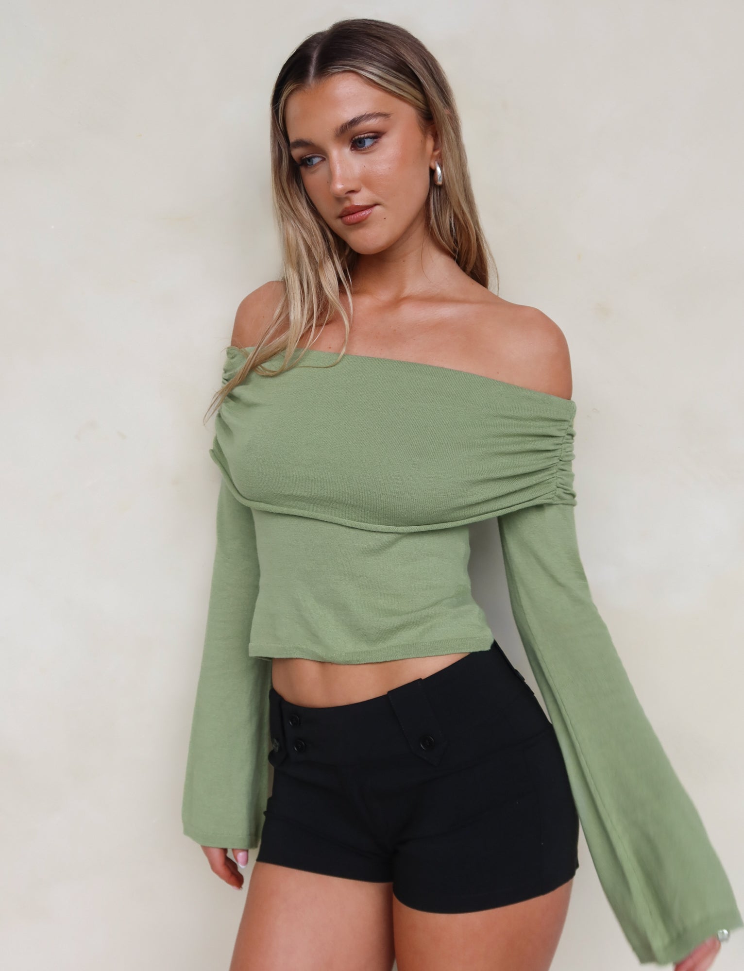 A woman with long, blonde hair is posing against an off-white background. She is wearing the DIEDRE JUMPER - GREEN from TIGERMIST, a light green, high stretch fabric, off-the-shoulder top paired with black high-waisted shorts. She has a neutral expression and her head is tilted slightly to the side.