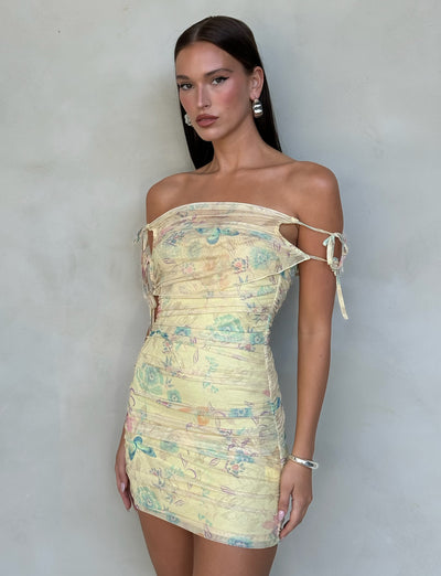 A woman with long, straight dark hair stands against a plain background. She is wearing the KERR DRESS - YELLOW : DELICATE FLORAL by TIGERMIST, an off-shoulder, form-fitting yellow mini dress with a subtle floral print and decorative ties on the sleeves. She accessorizes with drop earrings and a bracelet.