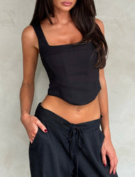 A woman with long dark hair is wearing the SULLIVAN TOP - BLACK by TIGERMIST paired with loose black drawstring pants. She poses with one hand in her pocket against a neutral background, highlighting her toned midsection and the relaxed yet stylish ensemble.
