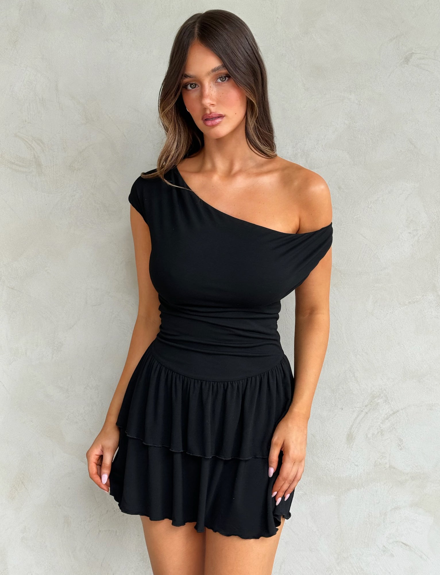 A woman with long brown hair is wearing the black IYANNA DRESS by TIGERMIST, an off-shoulder dress featuring a ruffled skirt. She stands against a textured, light gray background and looks directly at the camera with a neutral expression.