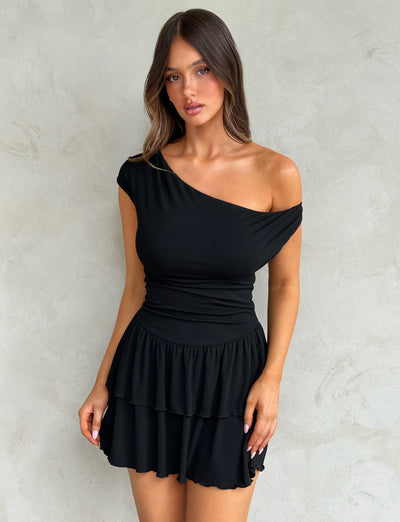 A woman with long brown hair is wearing the black IYANNA DRESS by TIGERMIST, an off-shoulder dress featuring a ruffled skirt. She stands against a textured, light gray background and looks directly at the camera with a neutral expression.