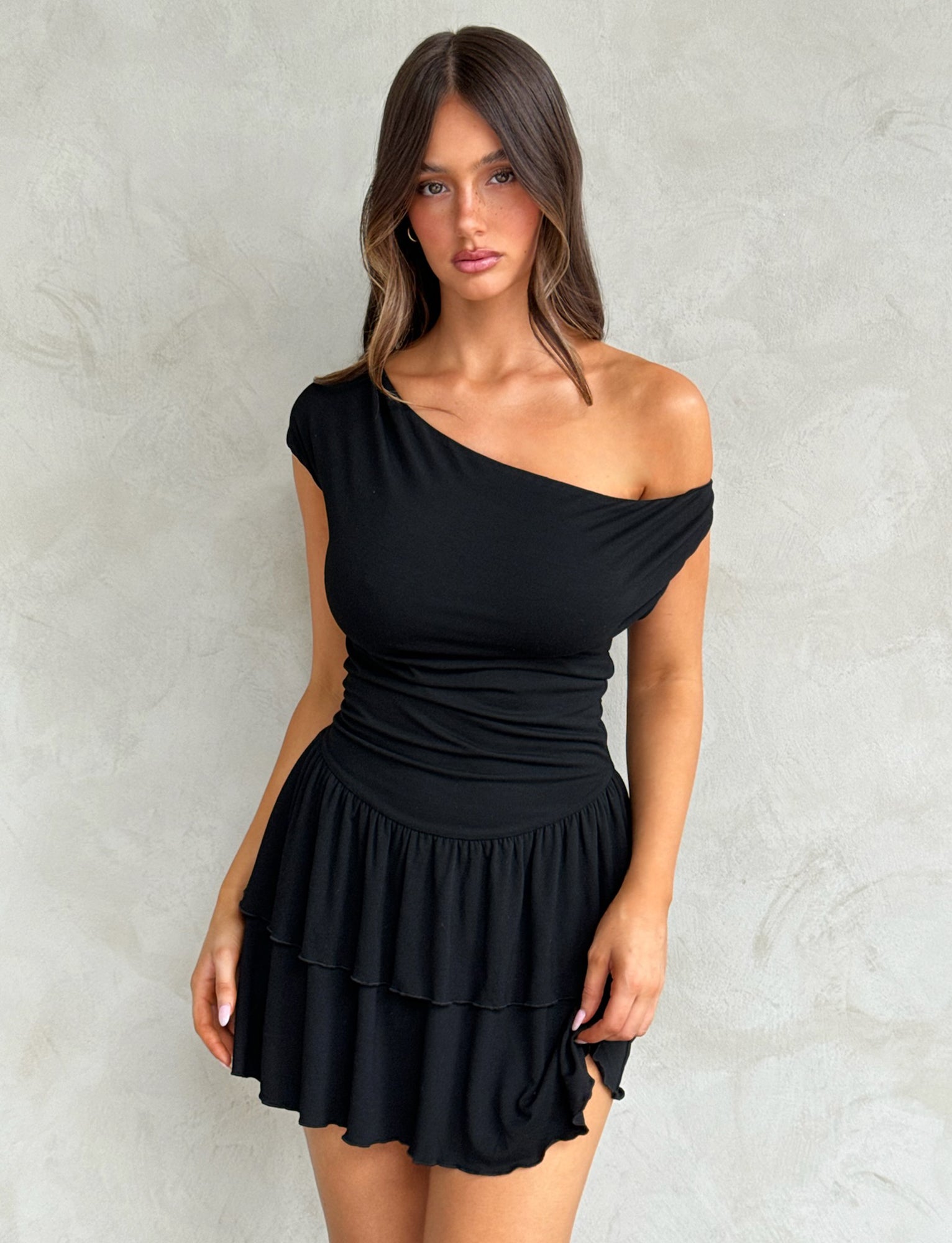 A woman with long brown hair stands against a textured gray background, wearing the IYANNA DRESS - BLACK from TIGERMIST. She gazes directly at the camera with a neutral expression, her arms hanging by her sides.