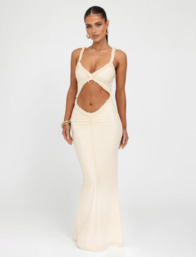 Wearing the RAYLA DRESS - WHITE : CREAM from TIGERMIST, a woman showcases its long, form-fitting silhouette with a unique midsection cutout design, spaghetti straps, and plunging neckline. Her long hair is pulled back as she accessorizes with hoop earrings and bangles, standing against a plain background.