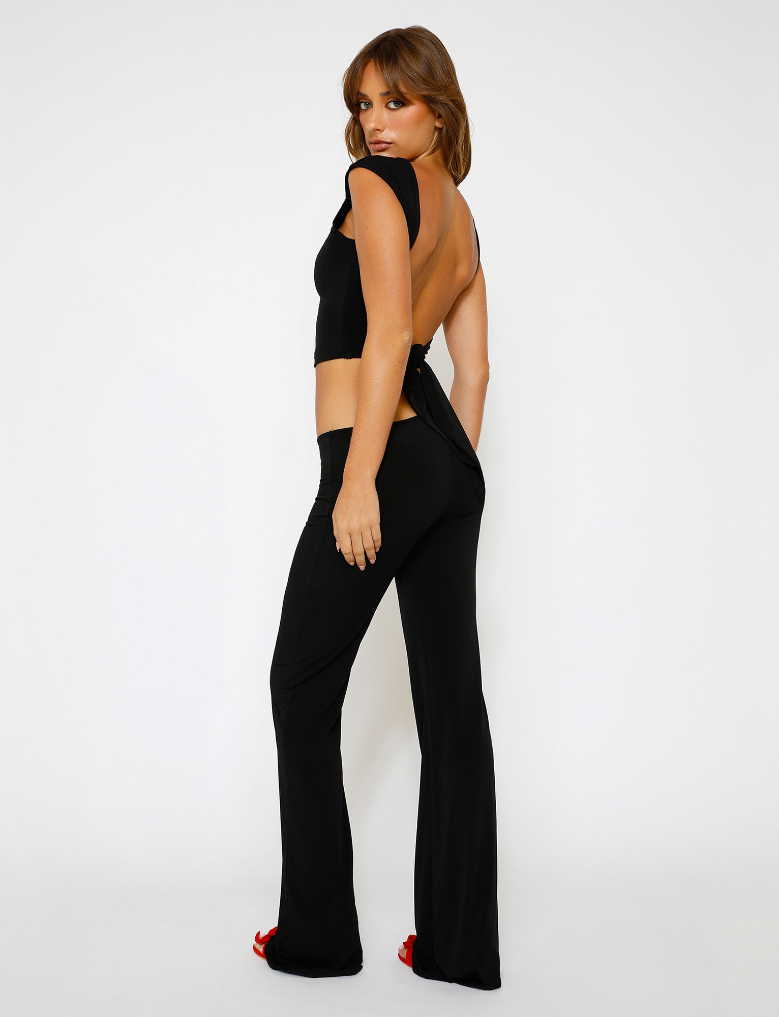 A woman stands against a plain white background, looking over her shoulder at the camera. She is wearing an open-back black top and the SHAYNA PANT - BLACK from TIGERMIST, with red high heels peeking out from the pant legs. Her brown hair is styled with loose waves.