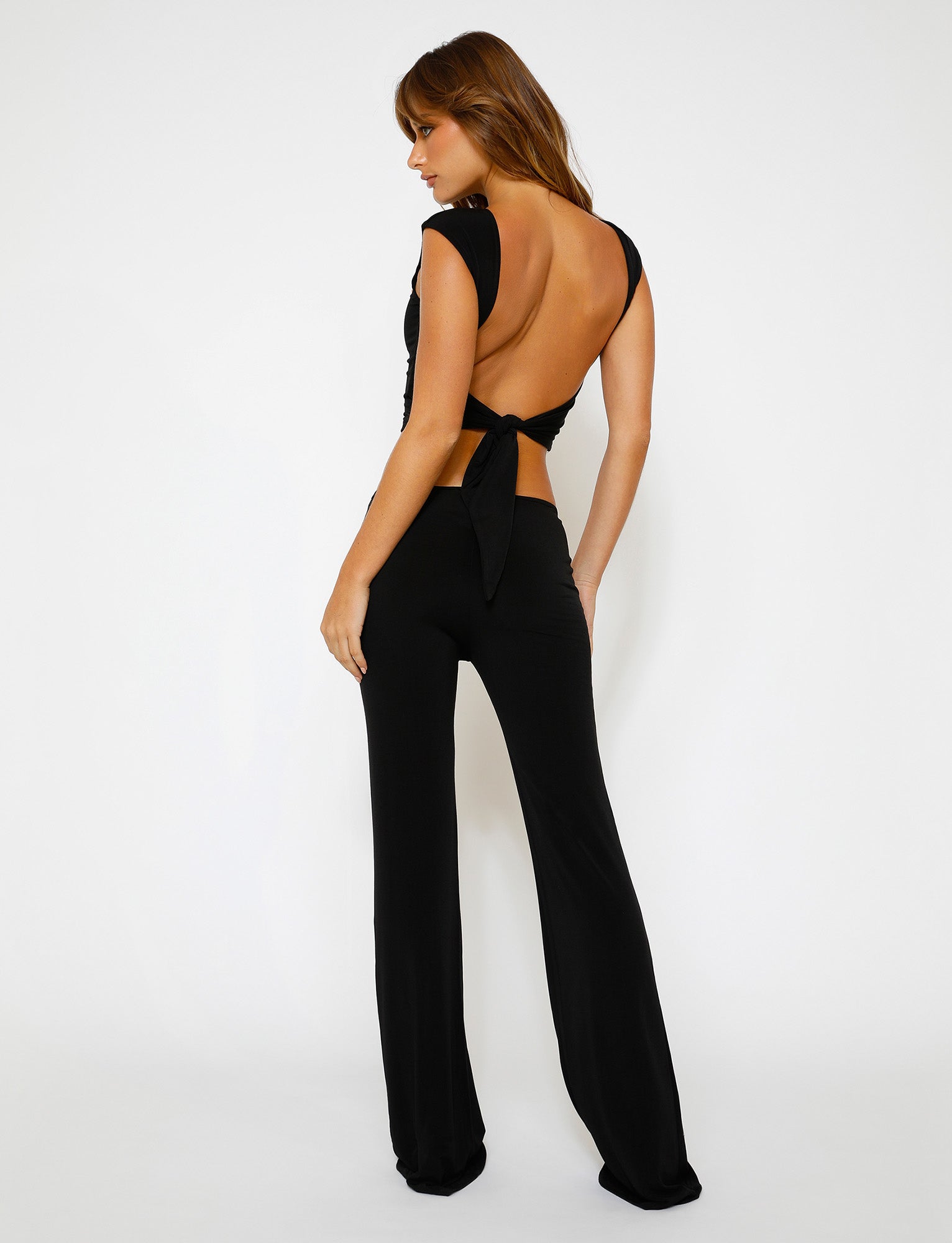 A person with long brown hair stands facing away, showcasing a backless black top with a tied bow paired with the SHAYNA PANT - BLACK from TIGERMIST. The outfit exudes an elegant yet daring style against a clean white background.
