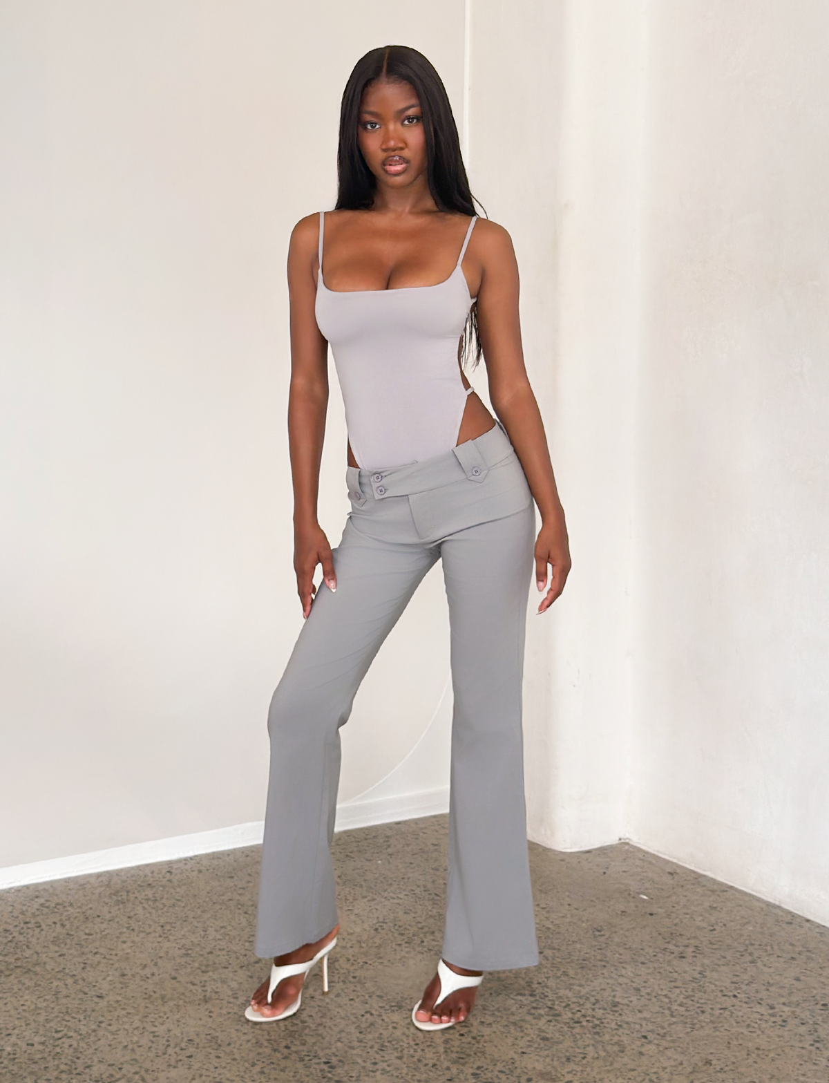 A woman stands indoors on a concrete floor, wearing TIGERMIST’s KITTIE PANT - FROST GREY and a fitted sleeveless top, paired with white heeled sandals against a plain light-colored wall.