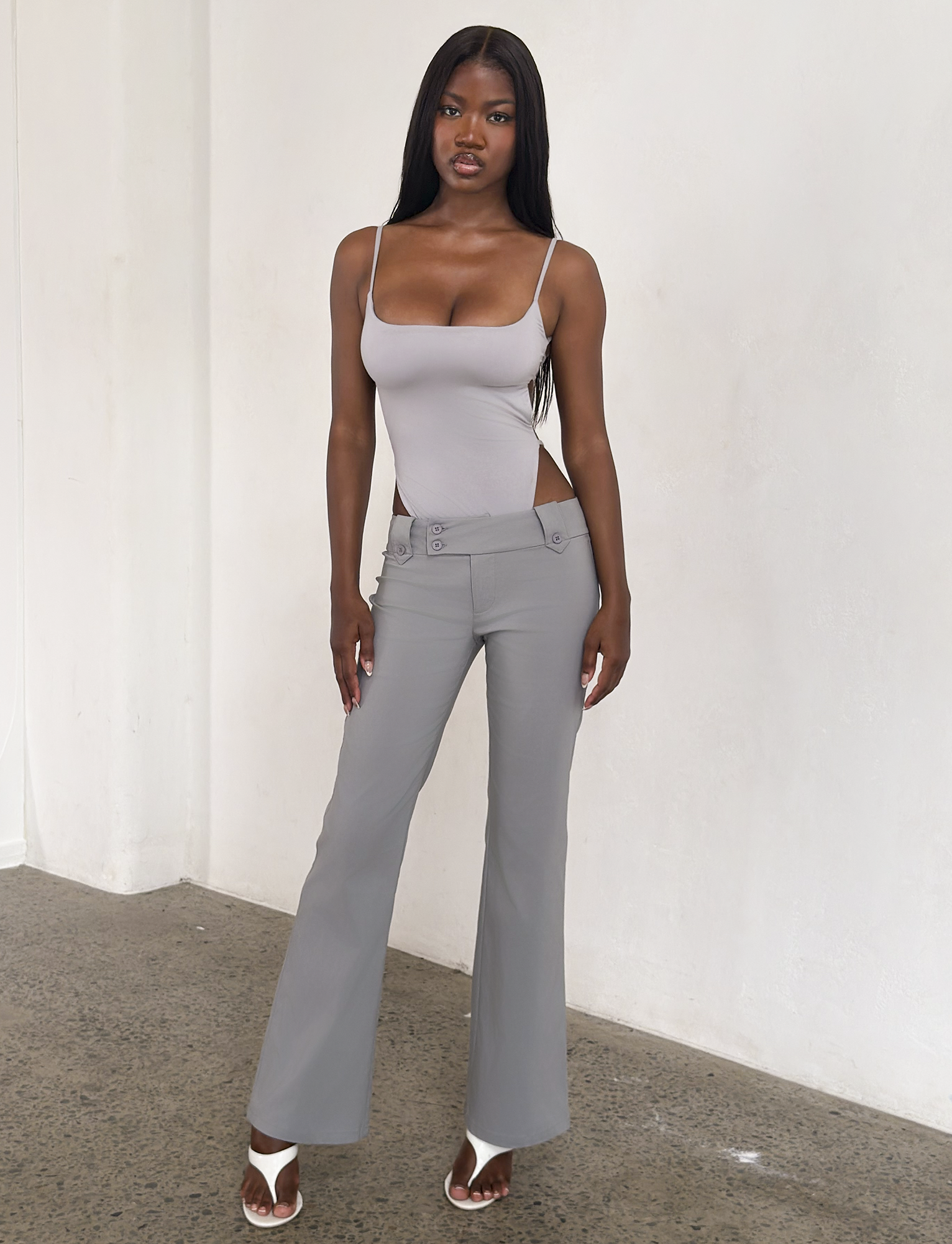 A woman stands indoors against a white wall, wearing a light gray tank, TIGERMIST KITTIE PANT - FROST GREY with cutout sides in stretch suiting fabric, and white heeled sandals. She has long dark hair and looks confident.