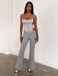 A woman stands indoors against a white wall, wearing a light gray tank, TIGERMIST KITTIE PANT - FROST GREY with cutout sides in stretch suiting fabric, and white heeled sandals. She has long dark hair and looks confident.