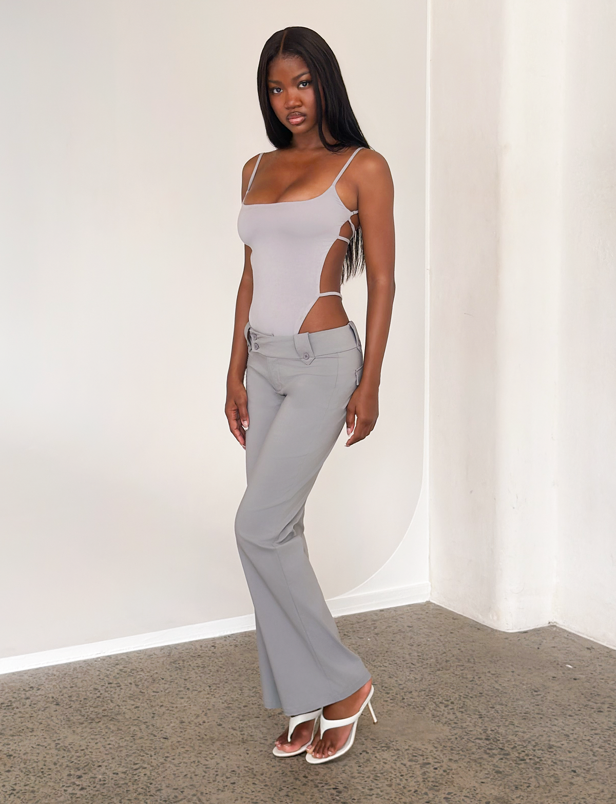 A woman poses indoors by a white wall wearing TIGERMIST's KITTIE PANT in frost grey and a light gray cut-out bodysuit, styled with white heeled sandals. She has long straight hair and stands on a concrete floor.