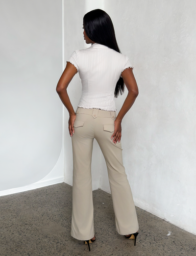 A woman with long black hair, wearing a short-sleeve white blouse and the TIGERMIST KITTIE PANT in sand stretch suiting fabric, stands in a minimalist room with light walls and a speckled floor, facing away.