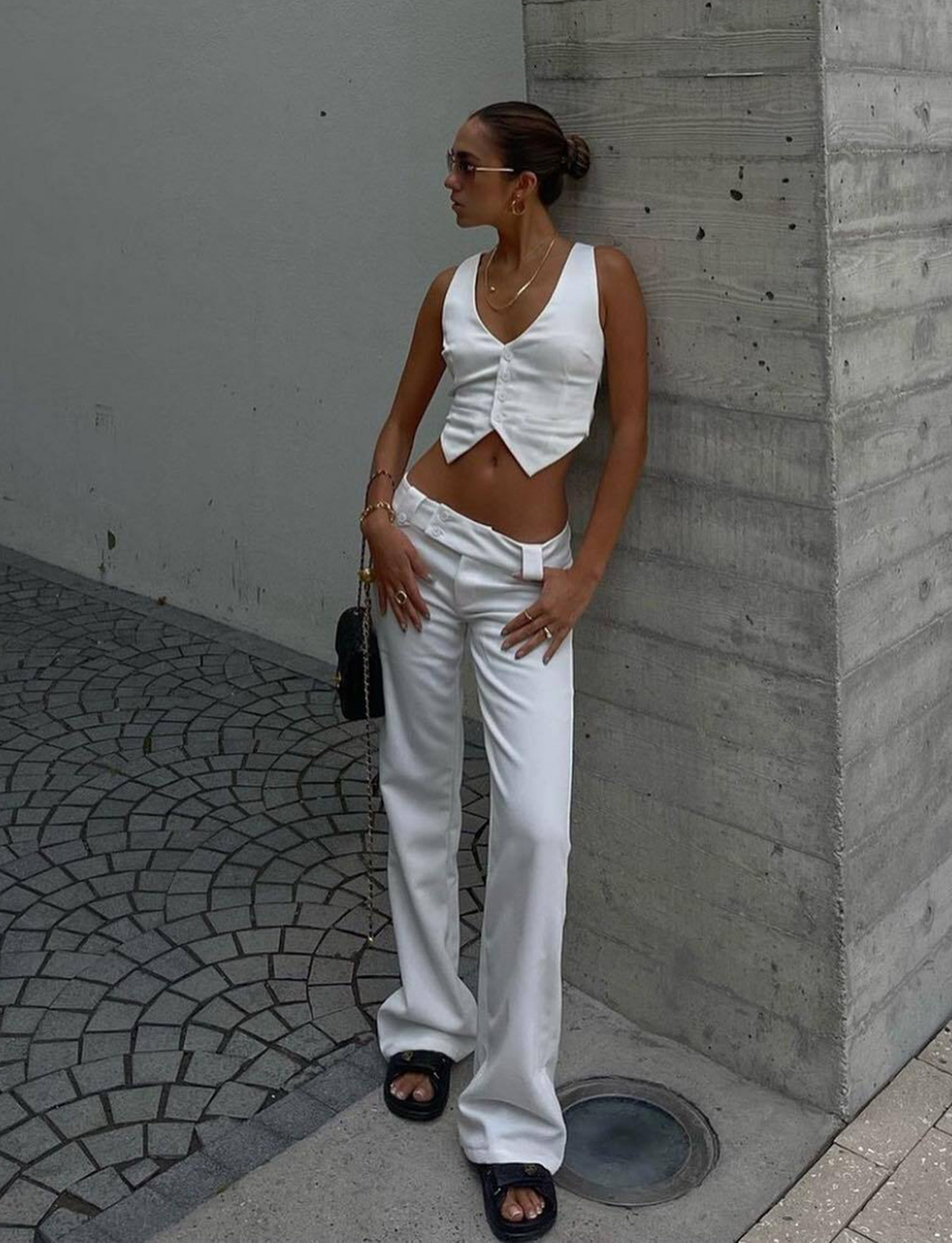 A woman stands outside against a concrete pillar, wearing a white cropped vest, TIGERMIST KITTIE TWILL PANT in white with a flared leg, black slide sandals, sunglasses, jewelry, and her hair styled in a sleek bun.