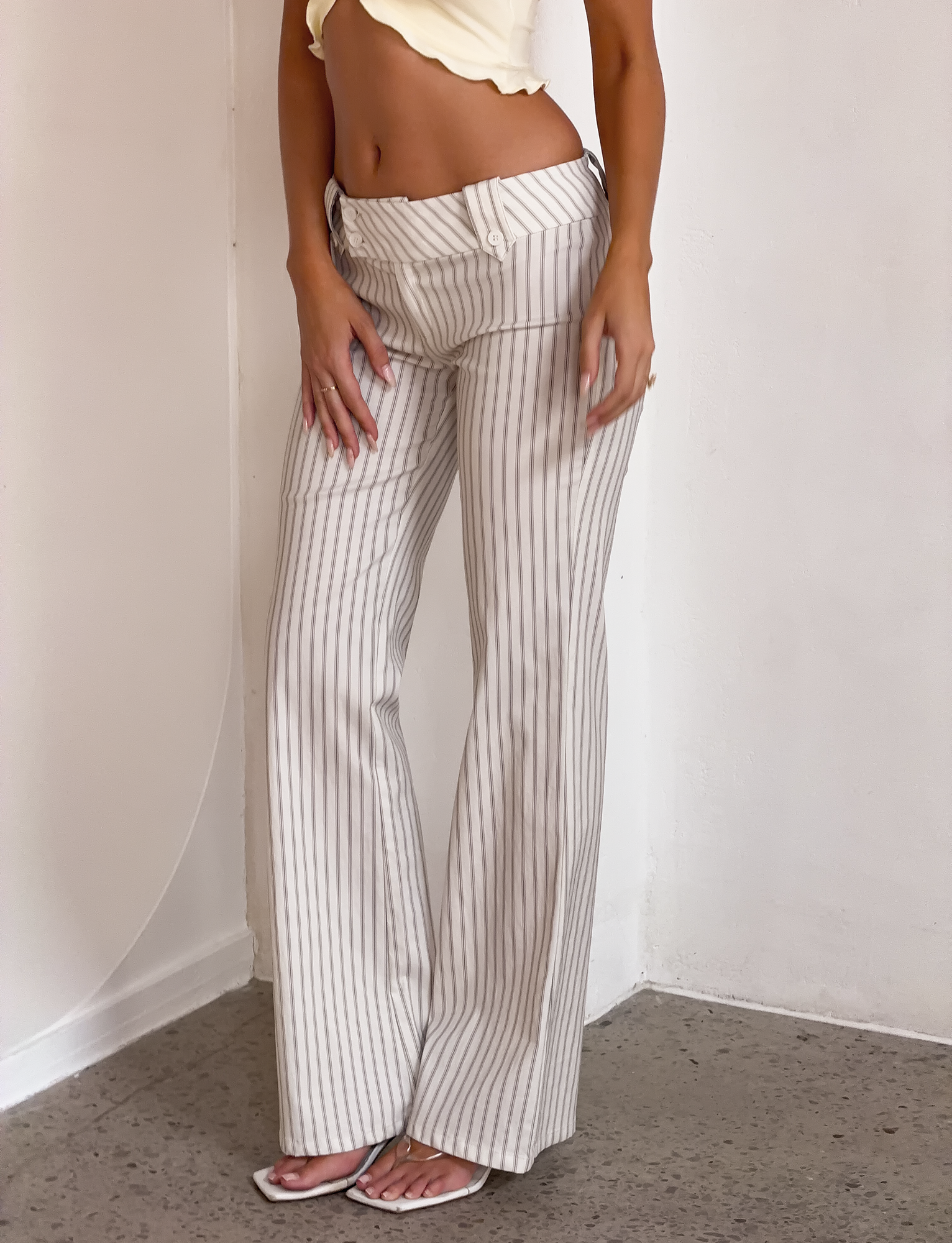 A woman in TIGERMIST KITTIE PANT - IVORY STRIPE, a high-waisted, wide-leg style with vertical gray stripes, pairs them with a cropped yellow top and white sandals, standing against a plain white wall.