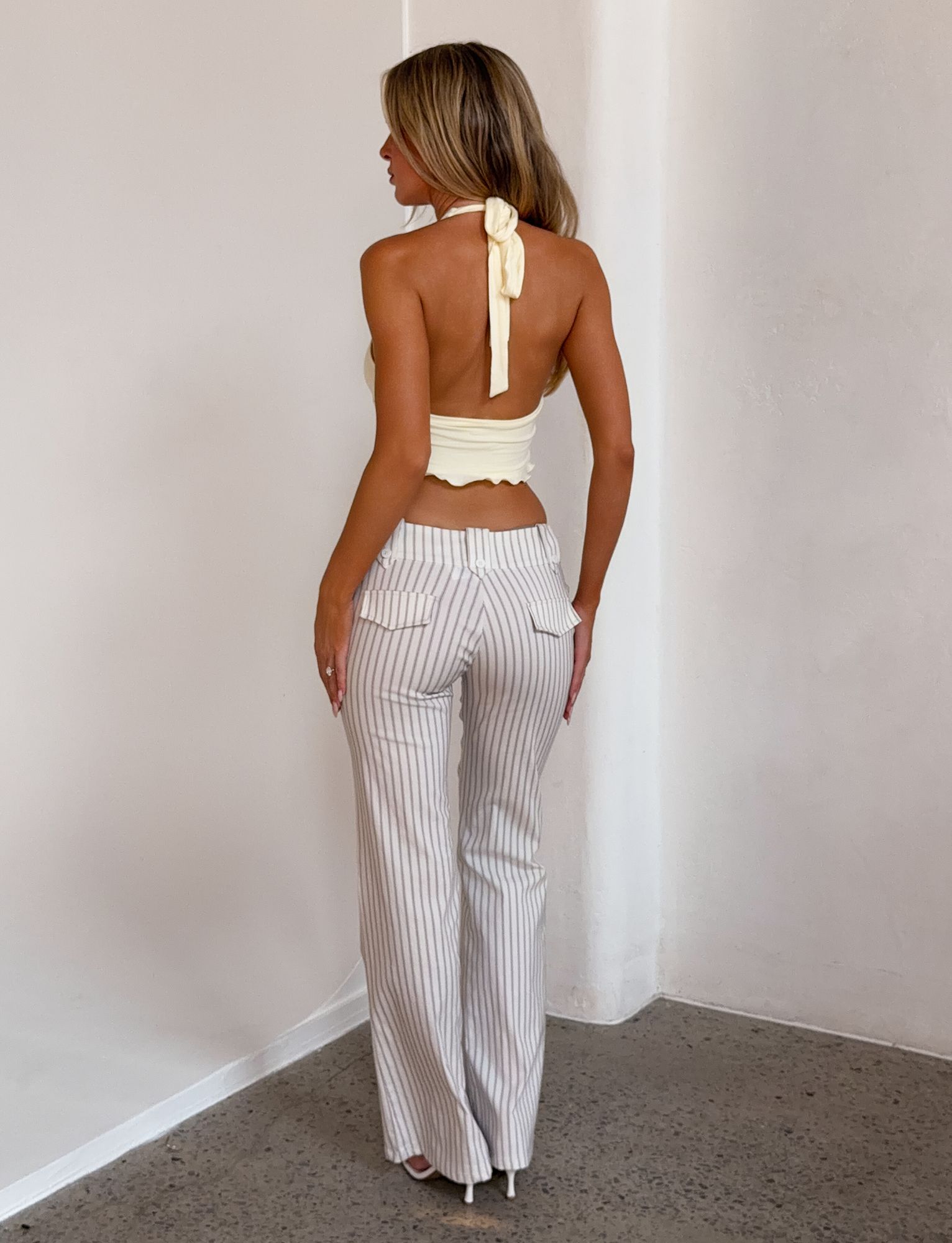 A woman with long, wavy hair stands indoors facing away, wearing TIGERMIST's KITTIE PANT - IVORY STRIPE, a light-colored, high-waisted flare style in stretch suiting fabric, paired with a yellow halter top and white heels on polished concrete.