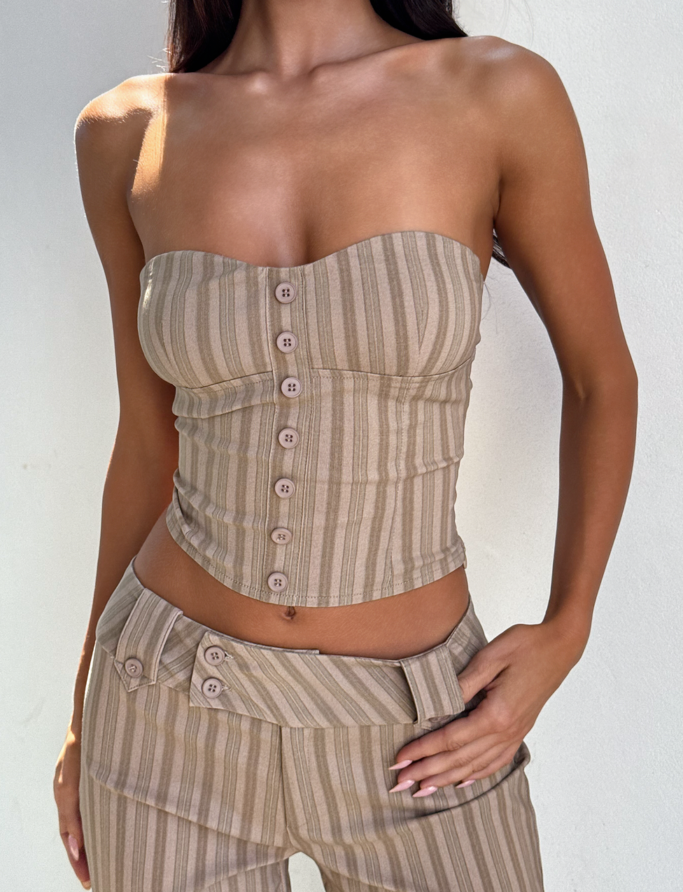 A woman wears the TIGERMIST KITTIE STRAPLESS CORSET in taupe stripe, featuring front buttons and paired with matching high-waisted stretch suiting pants. She poses confidently with one hand on her hip against a light background.