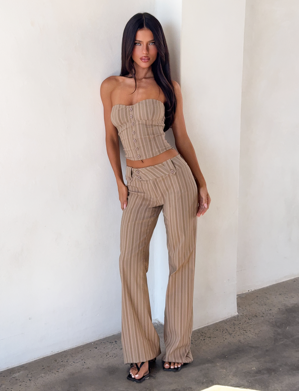 A woman with long dark hair poses against a white wall, wearing a strapless beige crop top and the TIGERMIST KITTIE PANT - TAUPE STRIPE in stretch suiting fabric, paired with sandals.