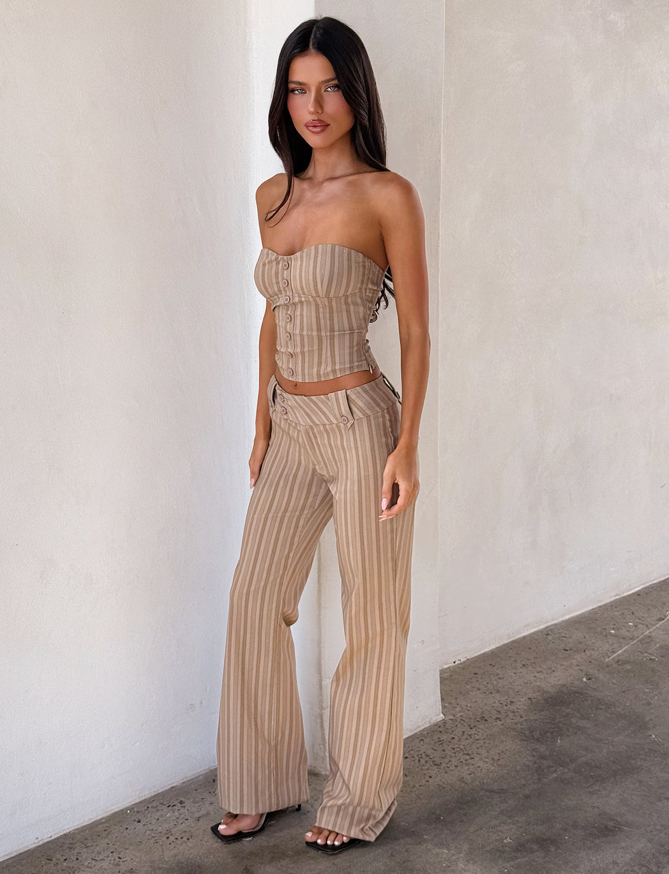 A woman with long dark hair stands indoors against a white wall, wearing TIGERMIST’s KITTIE PANT - TAUPE STRIPE in stretch suiting fabric and a matching strapless top, styled with black open-toe heels.