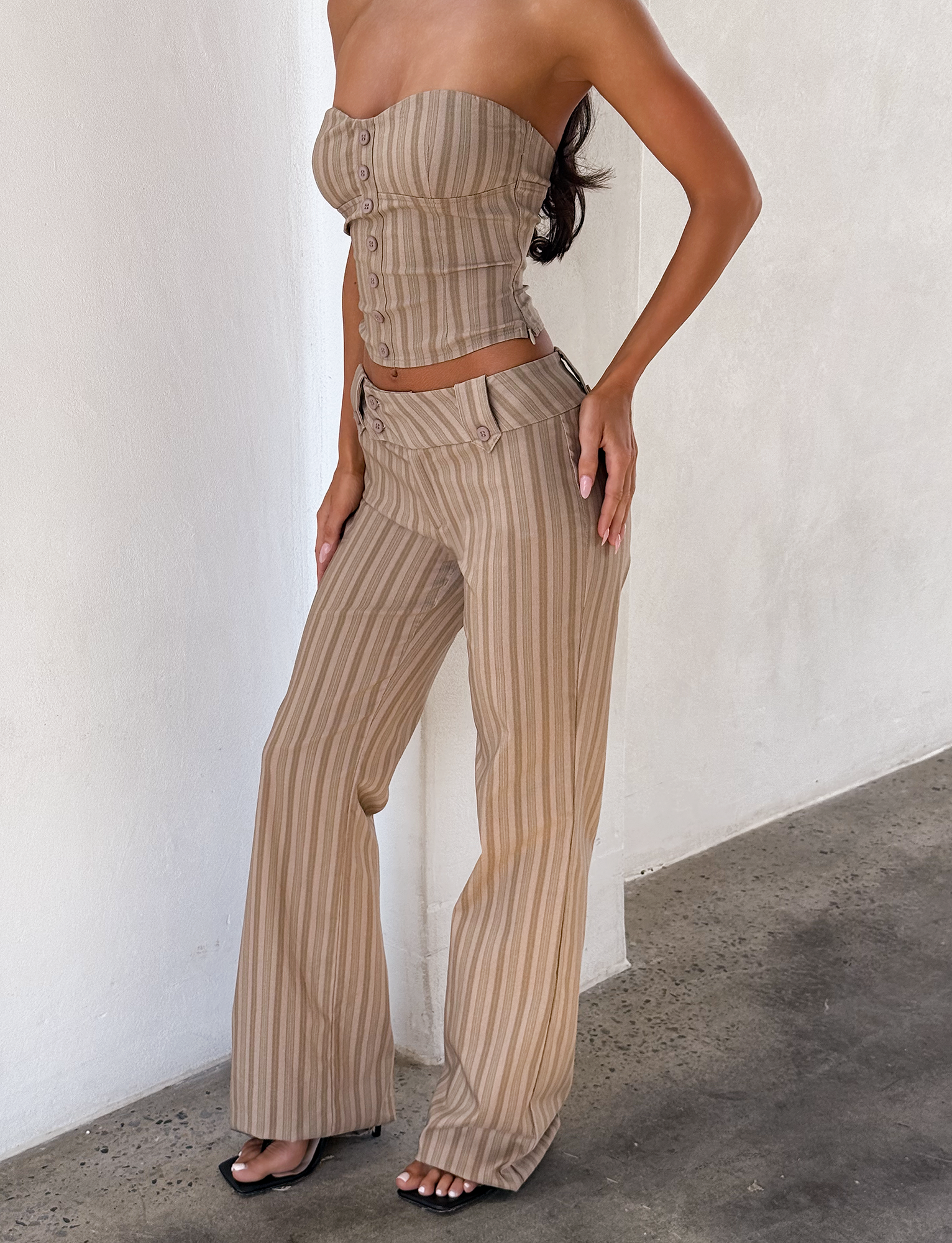 A woman wears a strapless, fitted, button-up crop top and TIGERMIST KITTIE PANT - TAUPE STRIPE, high-waisted wide-leg pants with vertical beige and taupe stripes, standing against a white wall on a concrete floor.
