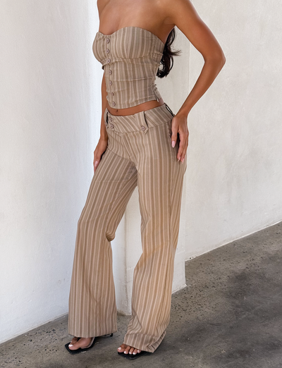 A woman wears a strapless, fitted, button-up crop top and TIGERMIST KITTIE PANT - TAUPE STRIPE, high-waisted wide-leg pants with vertical beige and taupe stripes, standing against a white wall on a concrete floor.