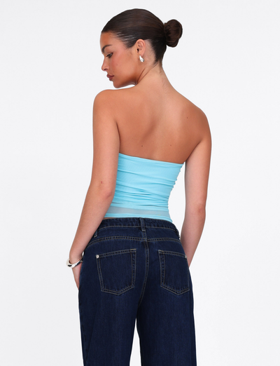 A woman with dark hair in a bun models the TIGERMIST HARRIETT TUBE TOP in ICE BLUE mesh and dark jeans, standing with her back to the camera, head turned slightly, accessorized with silver hoop earrings and a bracelet.