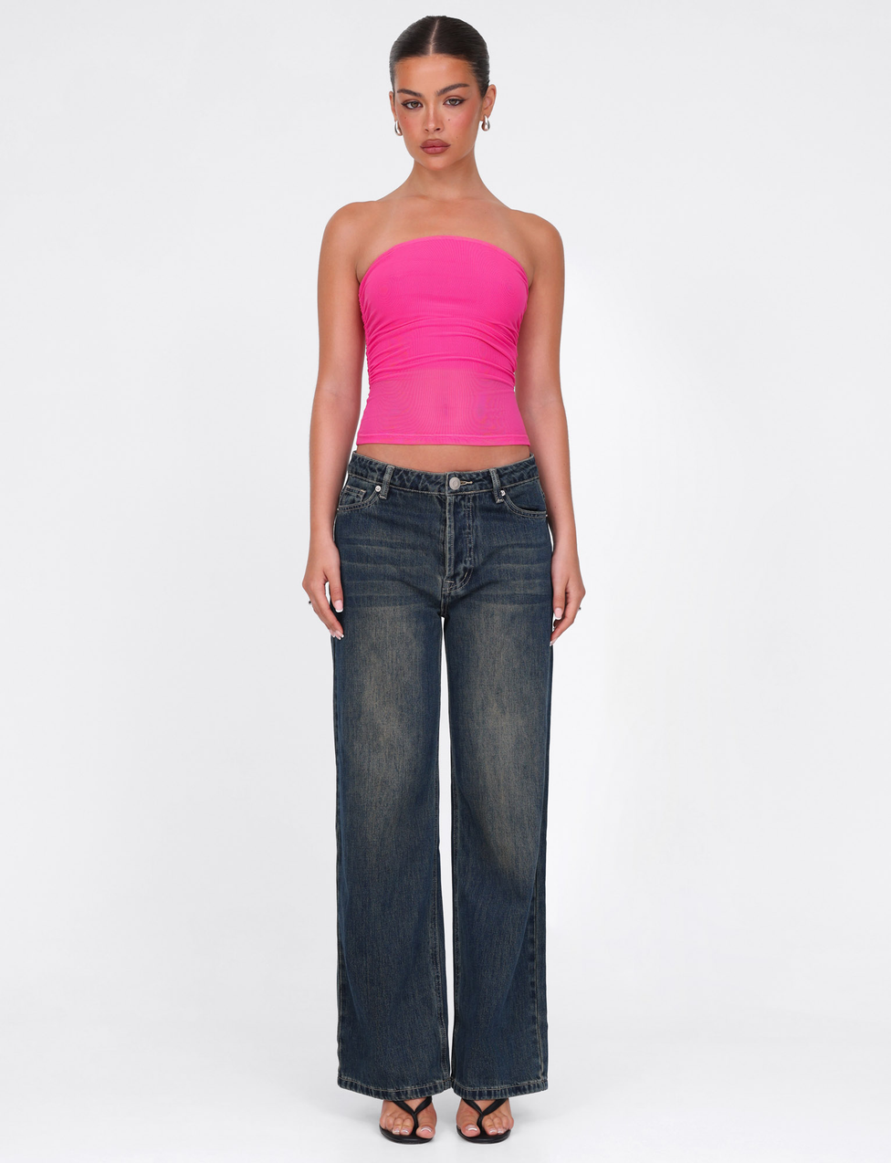 Against a plain white background, a person wears the TIGERMIST HARRIETT TUBE TOP in pink with wide-leg dark blue jeans, black heeled sandals, and hoop earrings. Their hair is tied back and their expression is neutral.