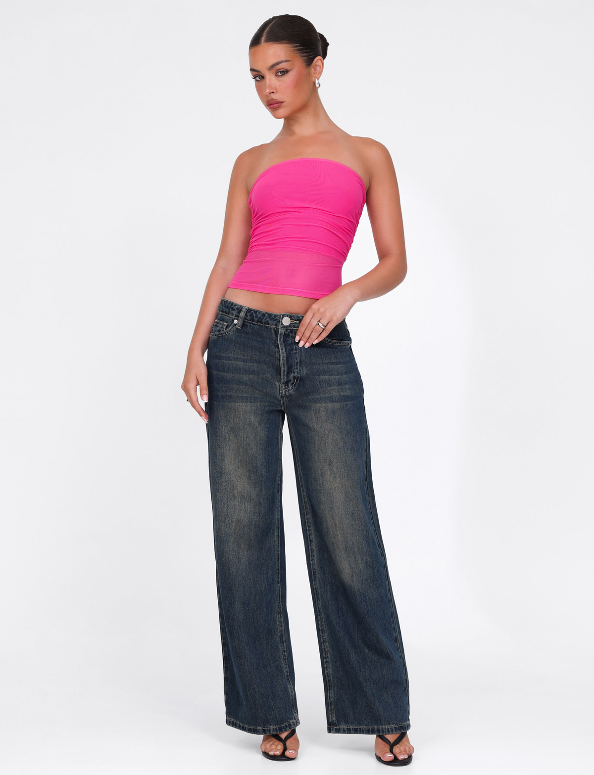 A woman poses with one hand on her hip in the bright pink TIGERMIST HARRIETT TUBE TOP, styled with high-waisted wide-leg blue jeans and black open-toe heels against a plain white background. Her hair is pulled back.