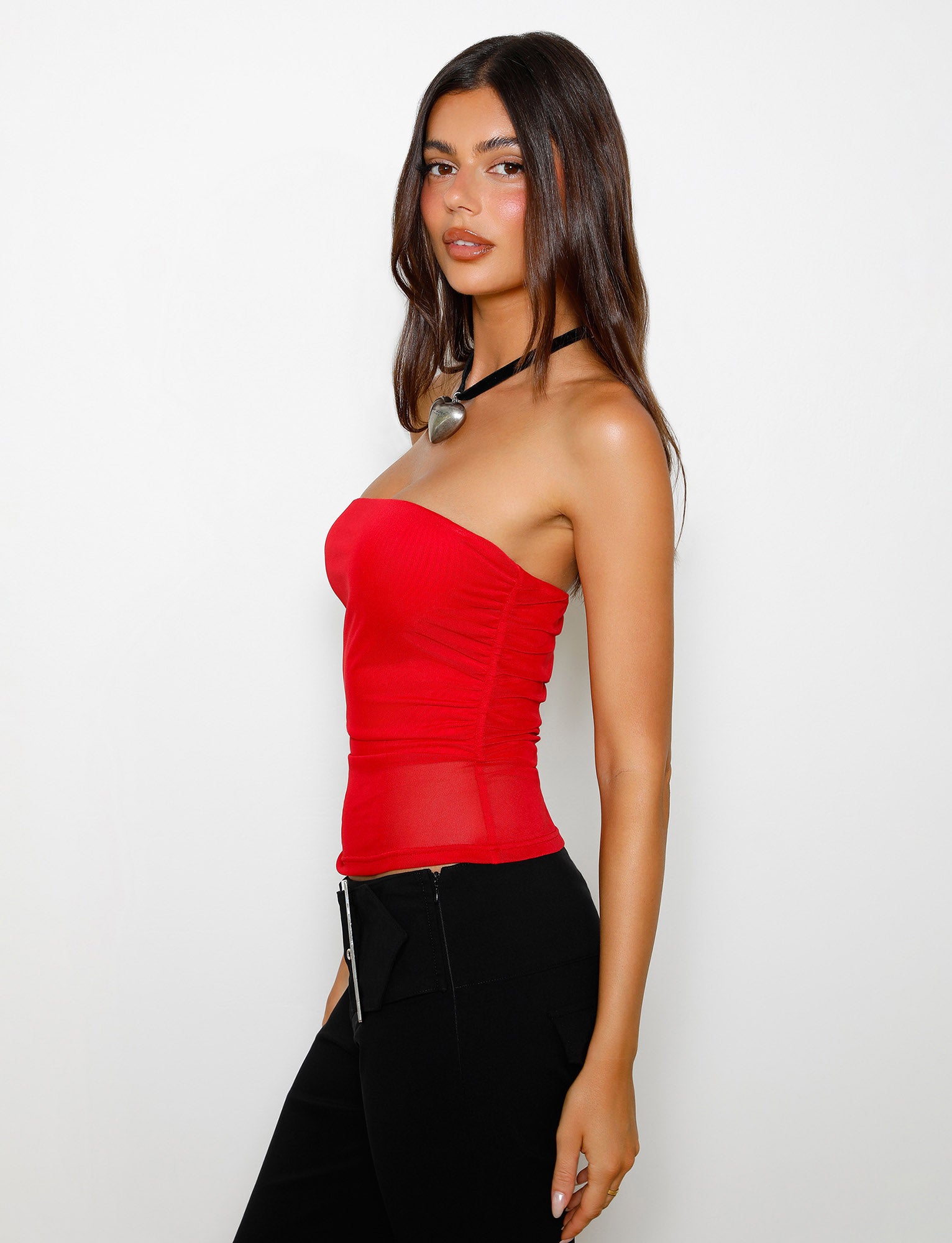A person with long dark hair is wearing the HARRIETT TUBE TOP - RED from TIGERMIST along with black pants. They are standing against a plain white background, looking at the camera with a neutral expression. In addition, they are accessorized with a thick black choker featuring a large pendant.