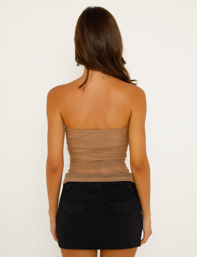 A woman with long brown hair is standing with her back to the camera. She is wearing the HARRIETT TUBE TOP - TAN from TIGERMIST and a short black skirt with pockets. The background is plain white.