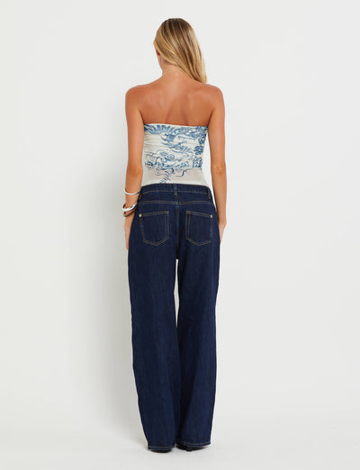 A woman with long blonde hair is shown from the back, wearing a strapless TIGERMIST HARRIETT TUBE TOP in blue with a tattoo print and high-waisted dark blue jeans. She has several bangles on her right wrist. The background is plain white.