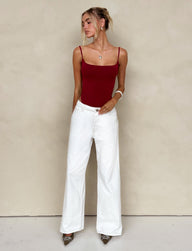 A woman stands against a beige wall wearing a TIGERMIST red spaghetti strap top and FELIX JEAN - WHITE wide-leg pants. One hand rests in her pocket while the other touches her arm. She has her hair styled in a casual updo and is accessorized with a necklace and bracelets, completing the look with pointed shoes.