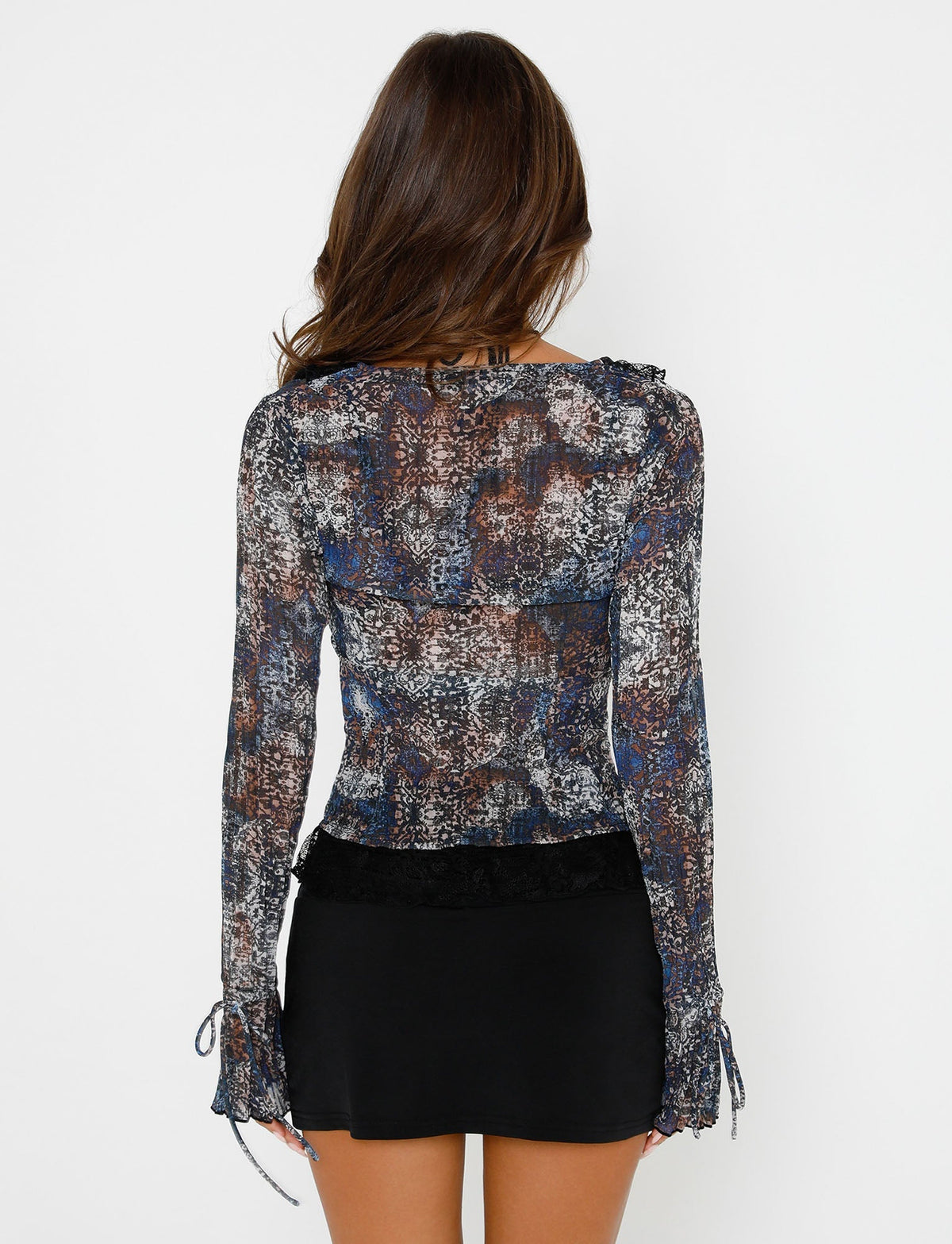 A woman with long brown hair stands facing away, showcasing the back of her TIGERMIST outfit. She is wearing the ZELDA TOP - BLACK ABSTRACT, a long-sleeved, semi-sheer top featuring a multicolored abstract pattern and ties on the sleeves. The top has a black lace band at the waist and is paired with a short black skirt.