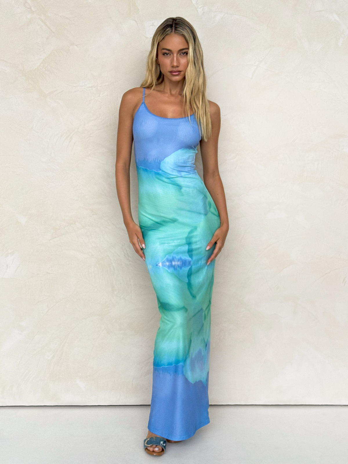 A woman with long blonde hair stands against a textured beige wall, wearing the TIGERMIST ARDON MAXI DRESS - BLUE : MACRO FLORAL : BLUE HIBISCUS, styled with blue heeled sandals.
