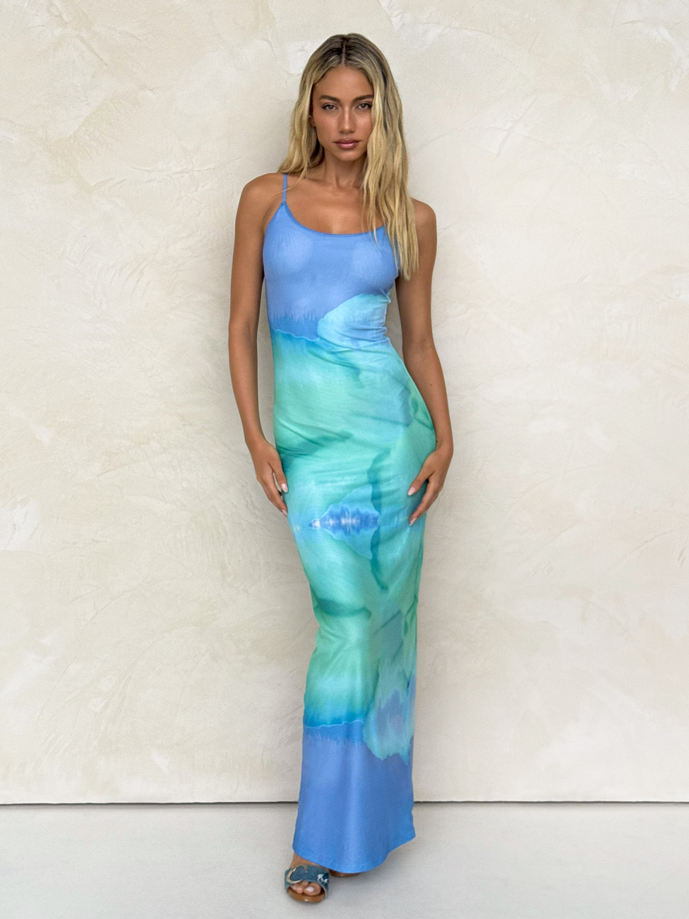 A woman with long blonde hair stands against a textured beige wall, wearing the TIGERMIST ARDON MAXI DRESS - BLUE : MACRO FLORAL : BLUE HIBISCUS, styled with blue heeled sandals.