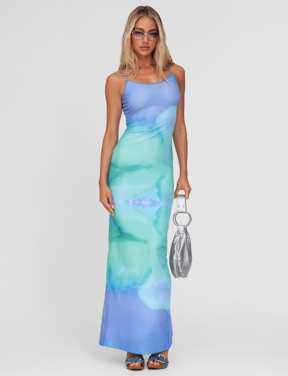 A woman stands against a white background wearing the TIGERMIST ARDON MAXI DRESS in Macro Floral Blue Hibiscus, blue sandals, sunglasses, and holding a silver handbag with a round handle.