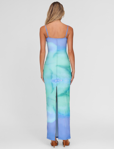 A woman with long blonde hair models the TIGERMIST ARDON MAXI DRESS in Blue Hibiscus Macro Floral, featuring thin straps and a back slit, standing against a plain white background with her back to the camera.