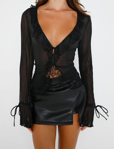 A person is adorned in a sheer black long-sleeved blouse with a deep neckline and lace details. The blouse, from Tiger Mist, is elegantly paired with the Daphnie PU Skort in black from TIGERMIST, which features a high waist and a small slit on the front. The background is plain white.