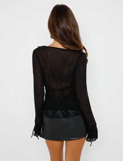 A woman with long brown hair stands facing away from the camera, wearing a sheer black long-sleeve top featuring textured vertical lines and lace detailing. She is also wearing the DAPHNIE PU SKORT - BLACK from TIGERMIST. The background is plain white.