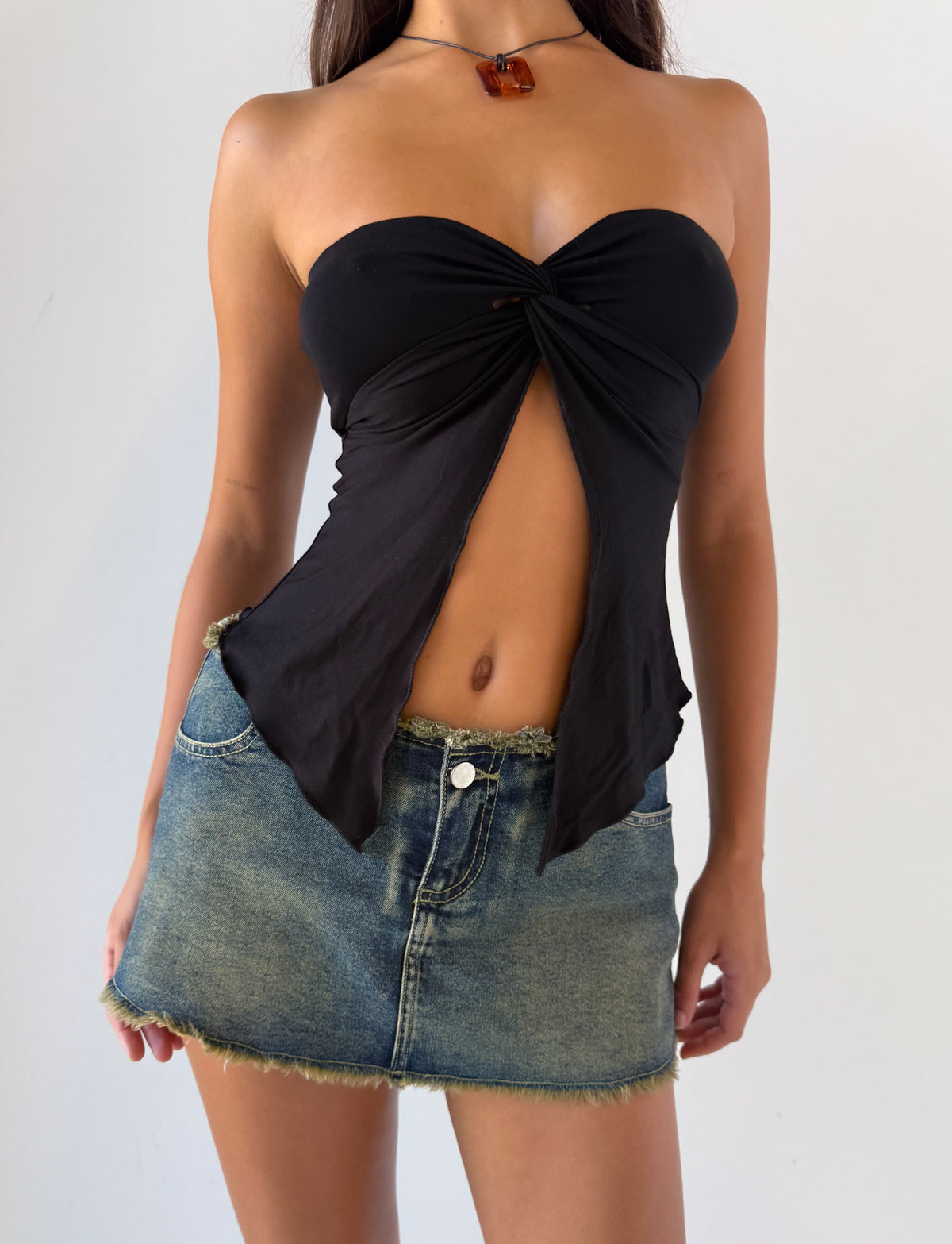 A woman wears the TIGERMIST CHARMAINE TOP in black, featuring a strapless knotted front, paired with a frayed denim mini skirt, standing against a plain white background.