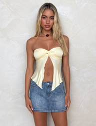 A woman with long blonde hair wears the TIGERMIST CHARMAINE TOP in Lemon and a blue denim mini skirt, standing against a textured light beige wall and looking directly at the camera with a neutral expression.