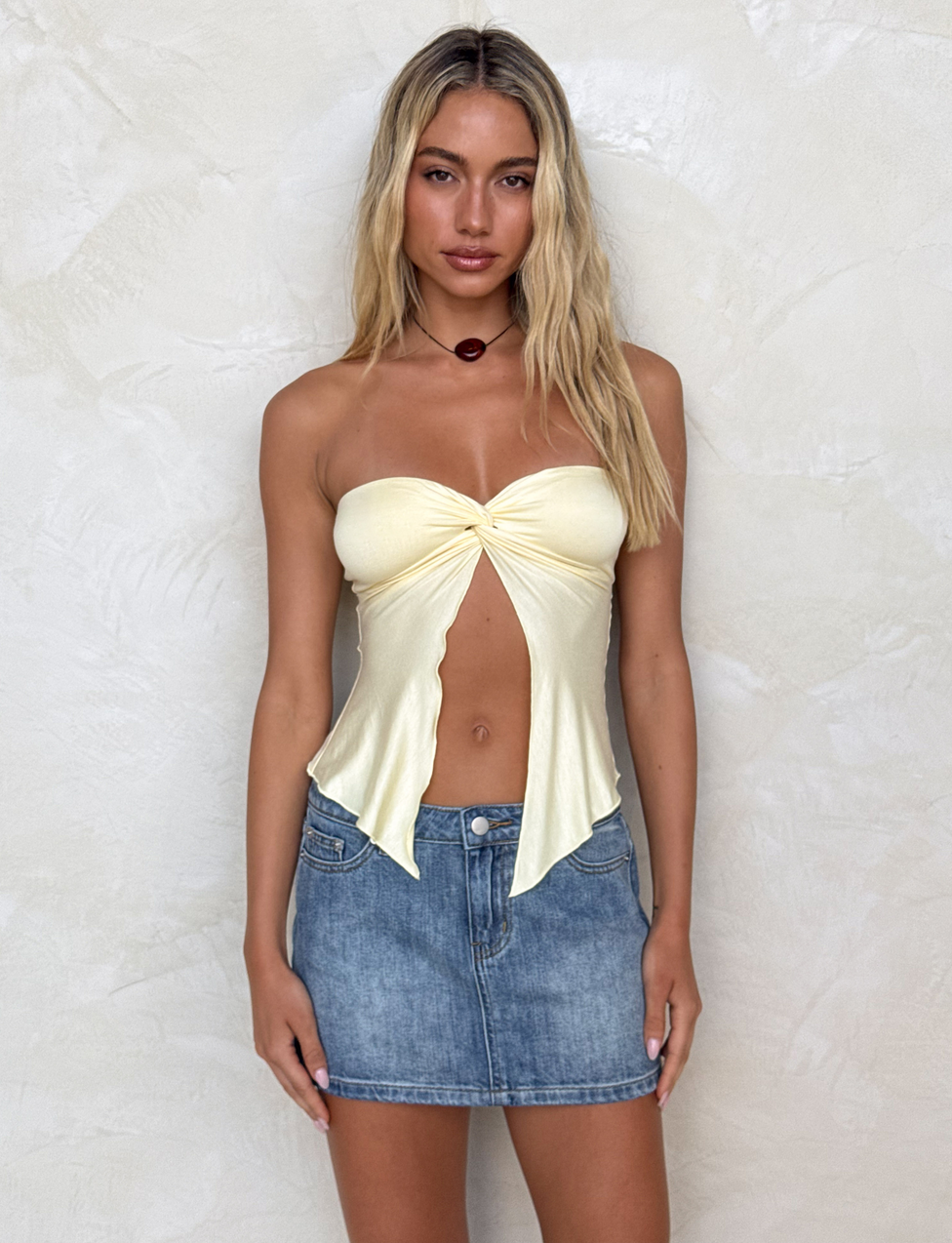 A woman with long blonde hair wears the TIGERMIST CHARMAINE TOP in Lemon and a blue denim mini skirt, standing against a textured light beige wall and looking directly at the camera with a neutral expression.