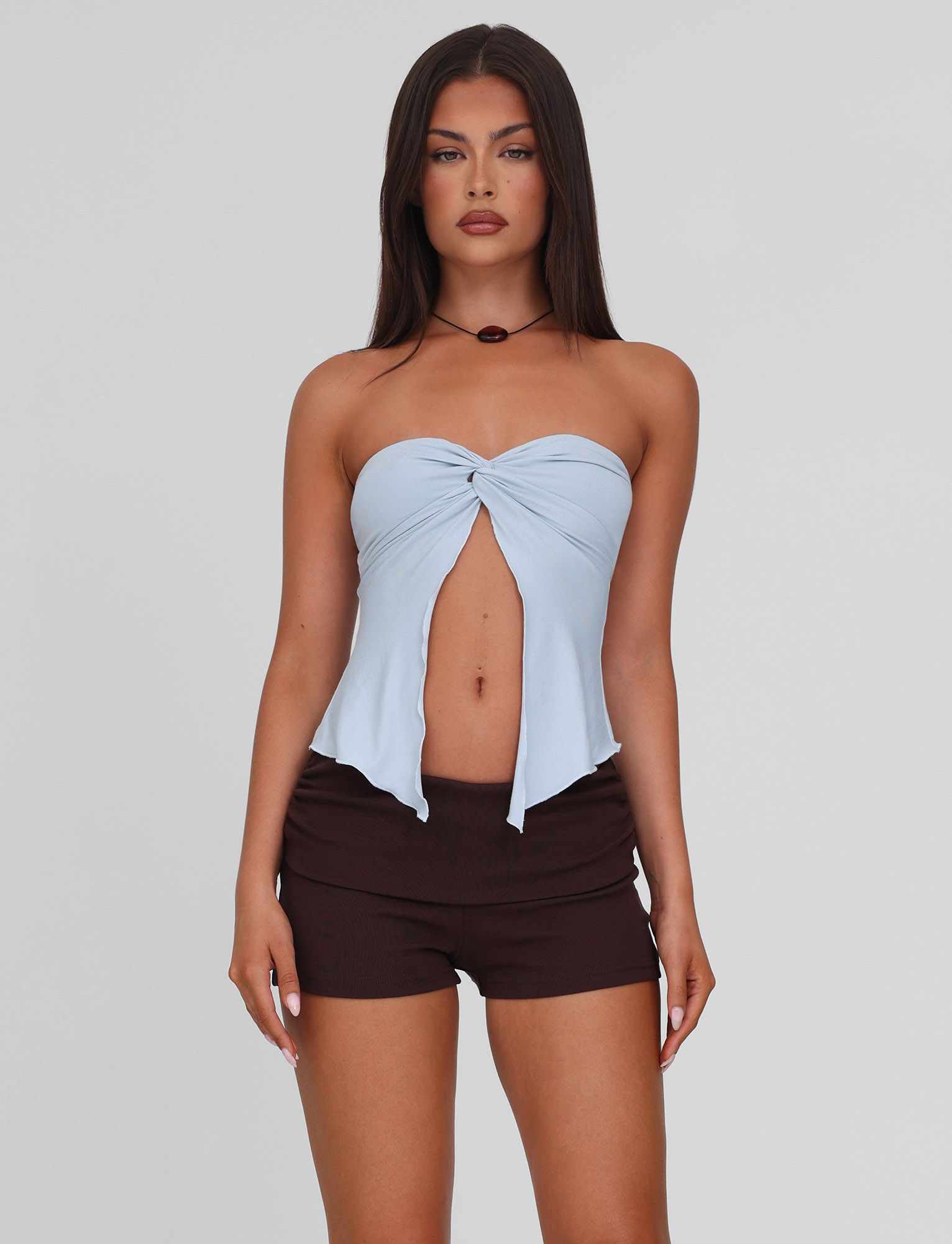 A woman with long dark hair wears the TIGERMIST CHARMAINE TOP in baby blue, featuring a strapless design with a front twist detail, paired with black shorts. She faces forward against a plain light gray background.