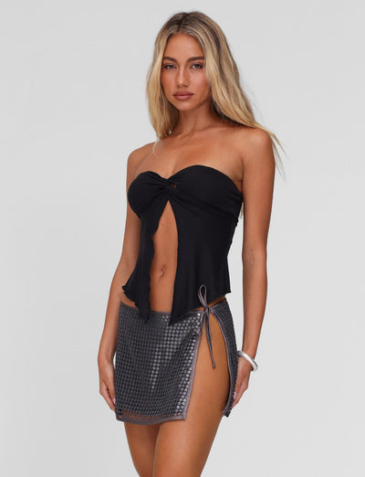 A woman with long blonde hair wears the TIGERMIST CHARMAINE TOP in black, a strapless, twist-detail top, paired with a short metallic-patterned skirt with a side tie, standing against a plain light background.